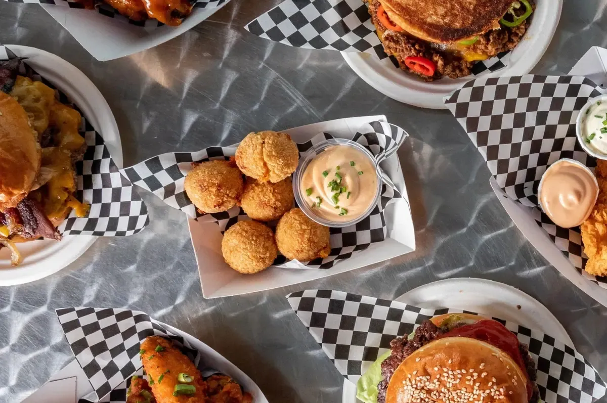 Hamburguesa de bacon en pan de sésamo sobre una bandeja de acero, servida en papel de charcutería a cuadros blancos y negros